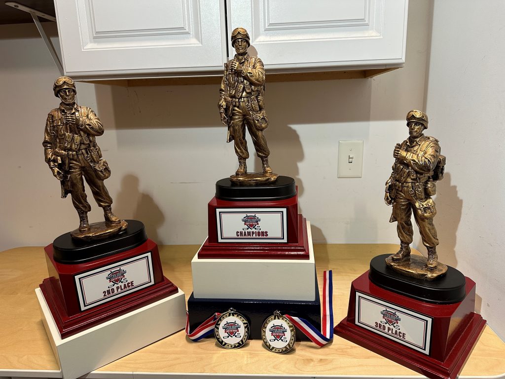 A picture of three trophies with second place on the left, first in the middle and third on the right. Two samples of the medals are in front of the first place trophy. all of the items are decorated with red, white and blue color scheme. The trophies are topped with a bronze soldier.